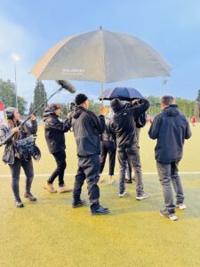 Eine Filmcrew in Regenjacken von hinten fotografiert. Ein Mann hält einen großen Schirm über das Equipment.