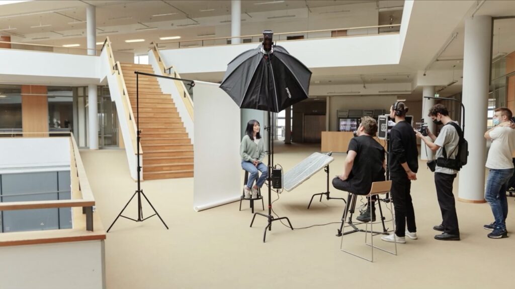 Eine freie Fläche in einem Bürogebäude, eine Frau sitzt vor einem weißen Hintergrund auf einem Hocker und wird interviewt und dabei gefilmt