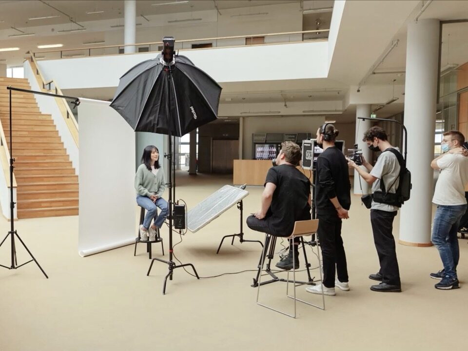 Eine freie Fläche in einem Bürogebäude, eine Frau sitzt vor einem weißen Hintergrund auf einem Hocker und wird interviewt und dabei gefilmt