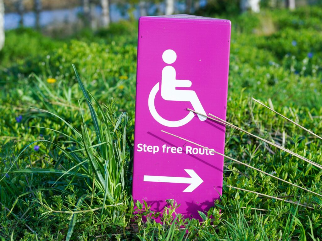 Ein pinkes Schild auf grünem Rasen auf dem ein Rollstuhlsymbol und der Text "Step free Route" zu sehen sind.